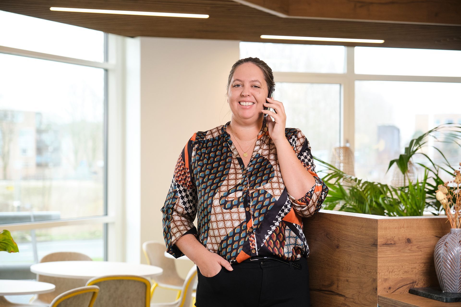 Een glimlachend persoon aan de telefoon in een lichte kantoorlounge naast een houten plantenbak.