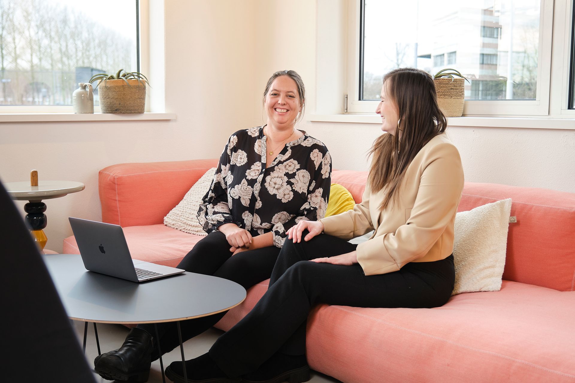 Twee mensen zitten te praten op een roze bank naast een laptop in een lichte kantoorlounge.