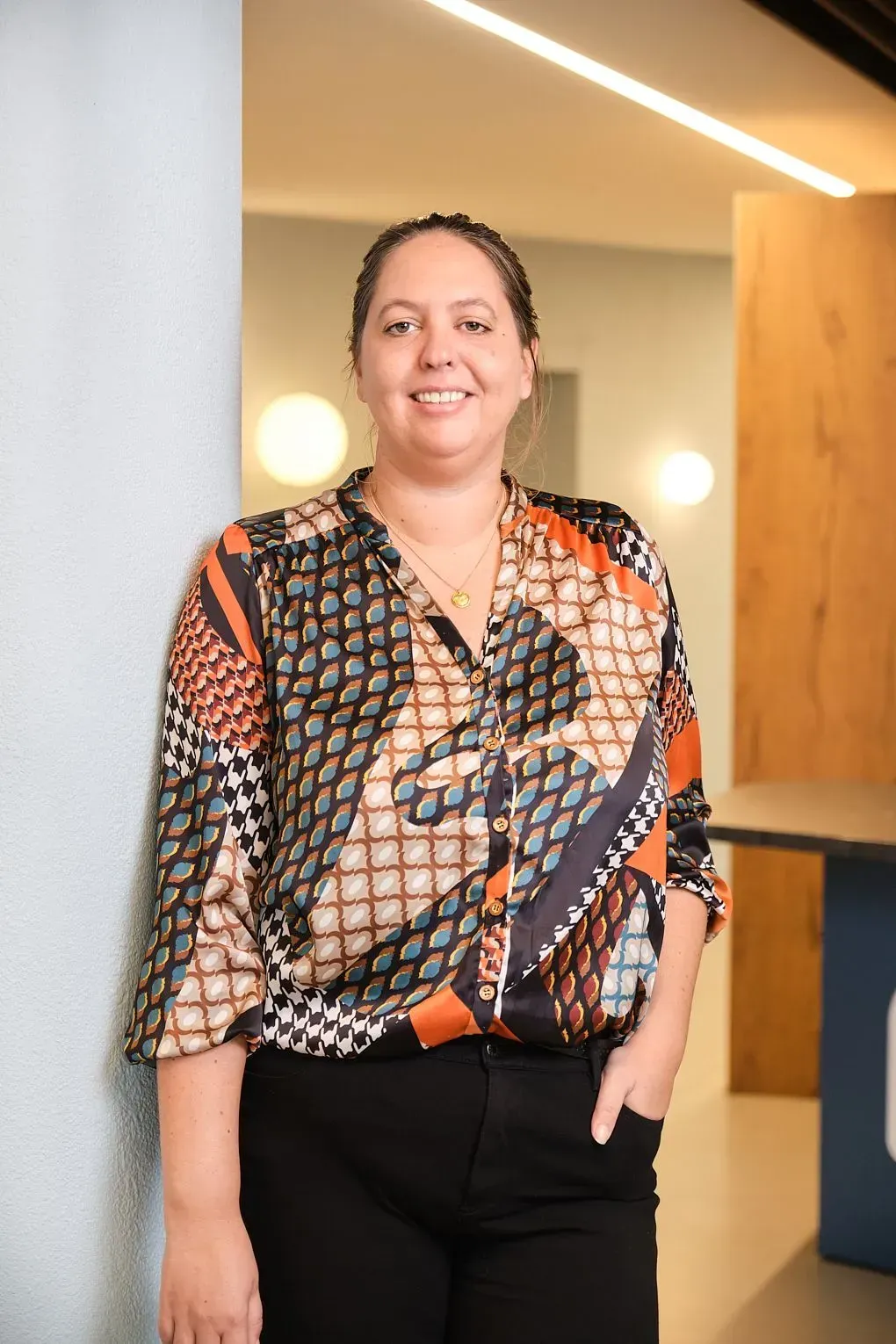 Vrouw staat lachend in een moderne hal, gekleed in een blouse met patroon en een zwarte broek.