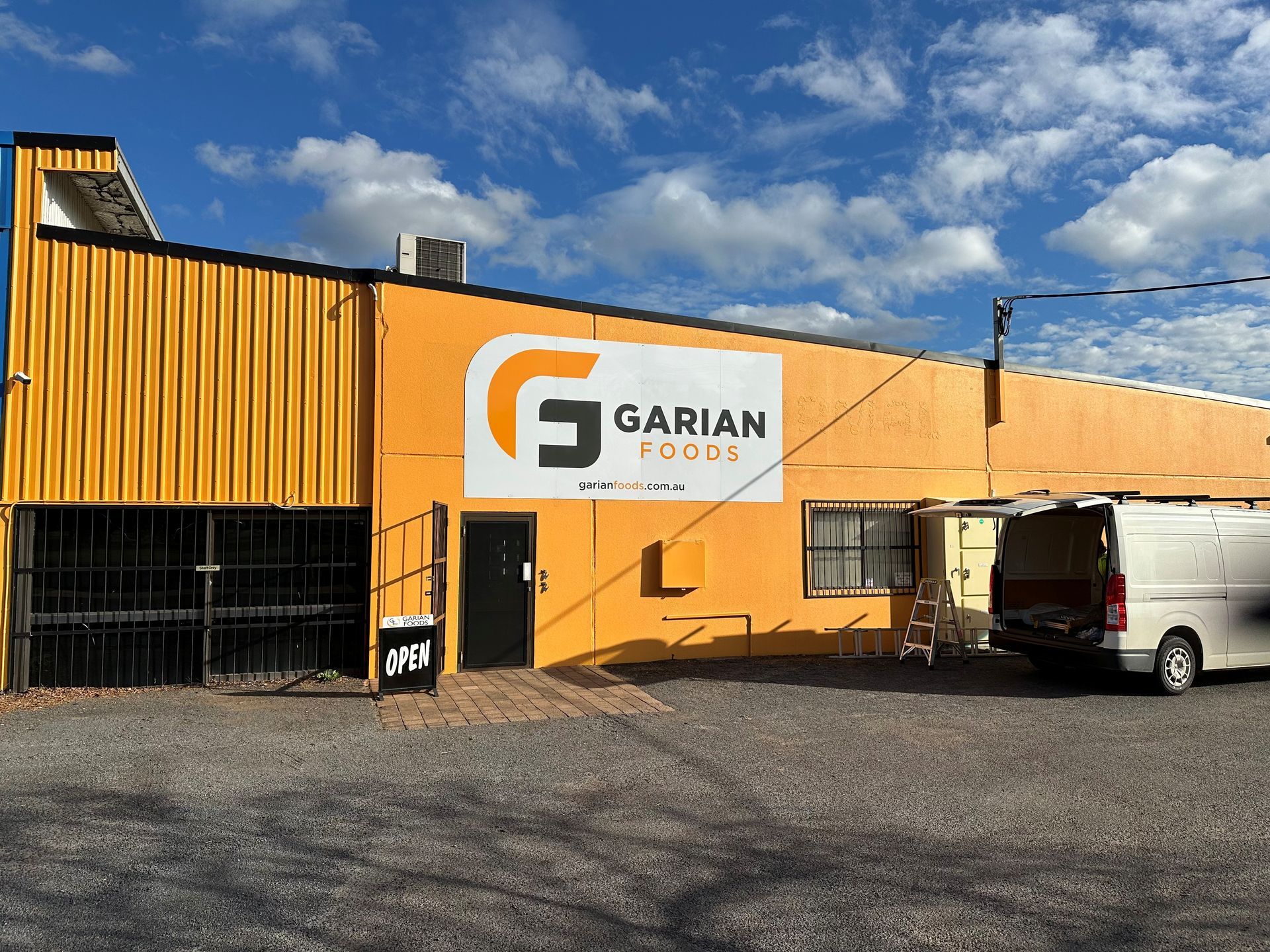 Yellow building with a Garian Foods sign, featuring a gravel parking area and a white van parked in front.