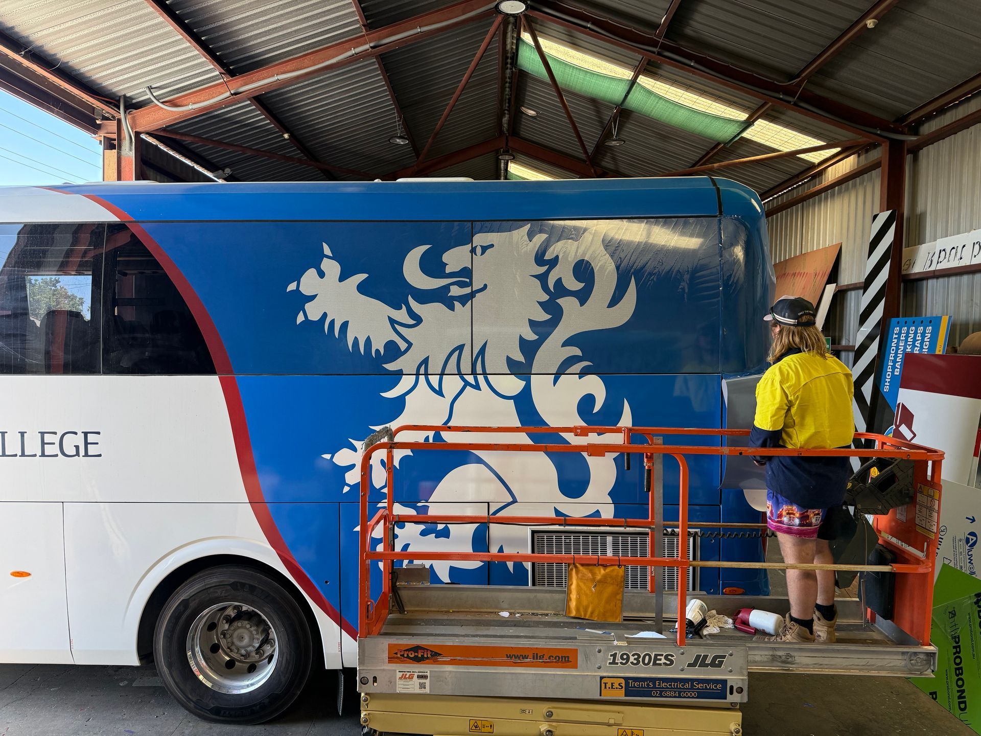 A person wearing a high-visibility yellow shirt works on a large blue and white lion graphic on the side of a bus.
