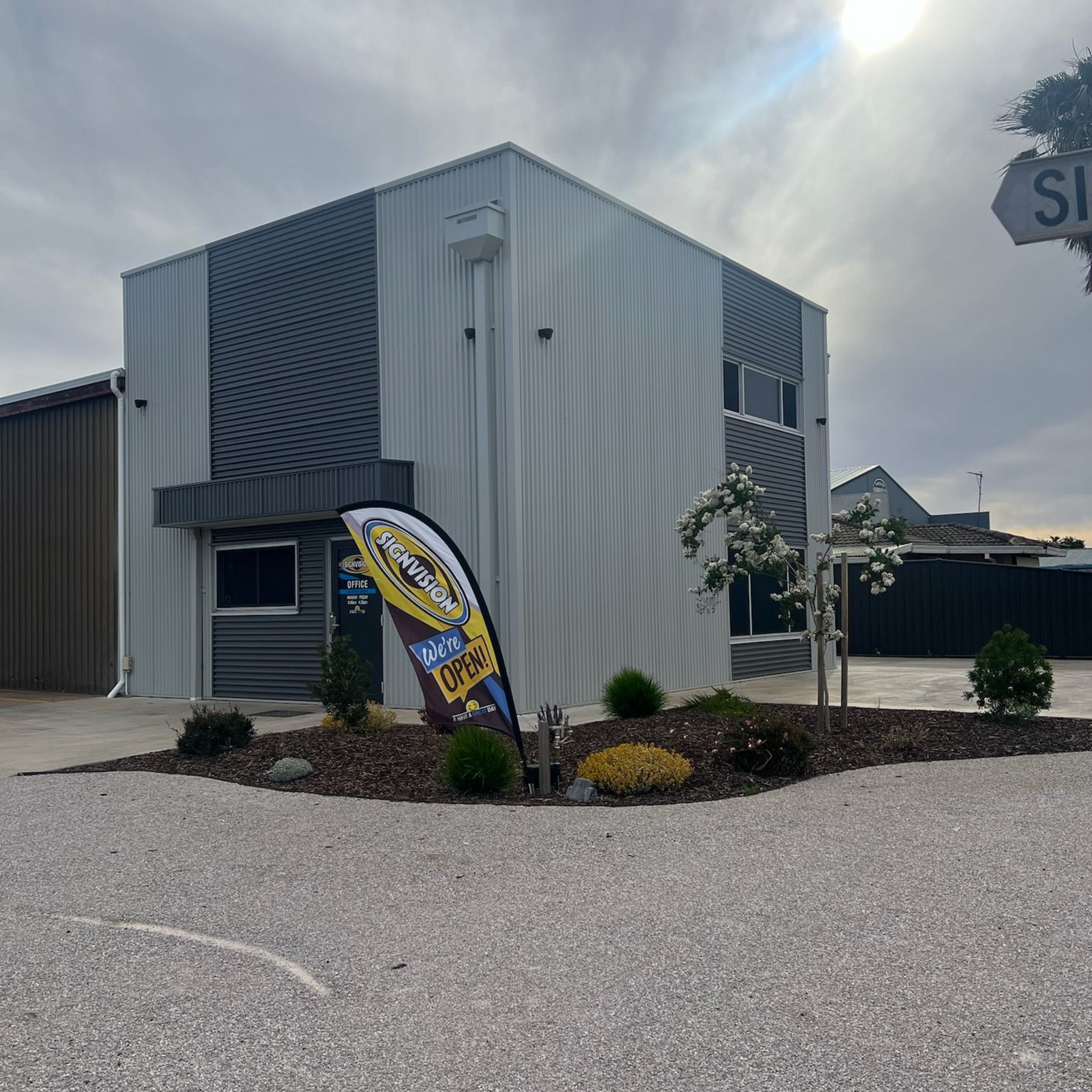 Modern industrial building with wavy gray panels and colorful sign. Gravel lot, cloudy sky.