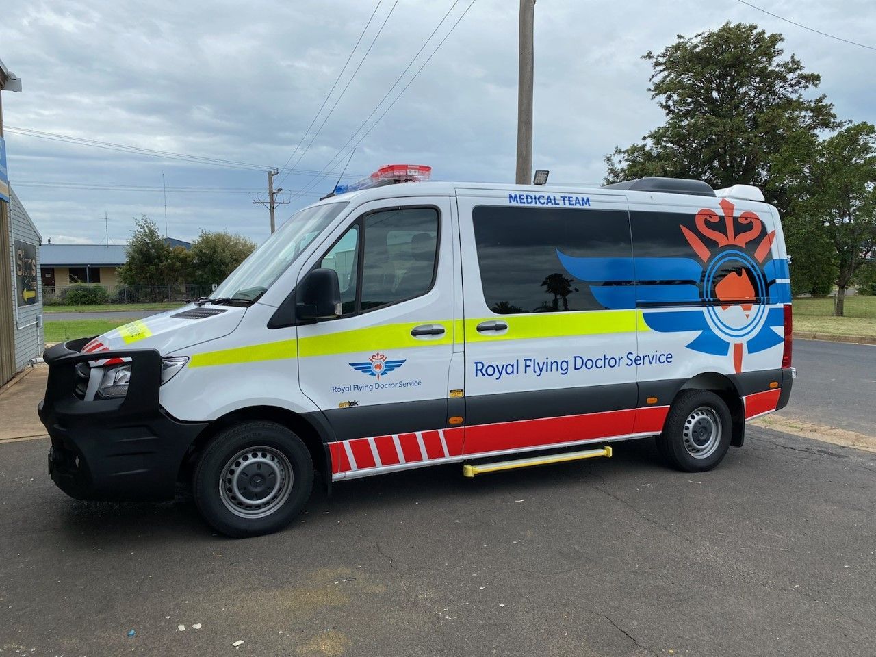 A white and red Royal Flying Doctor Service van with yellow markings, parked on an asphalt lot under a cloudy sky.