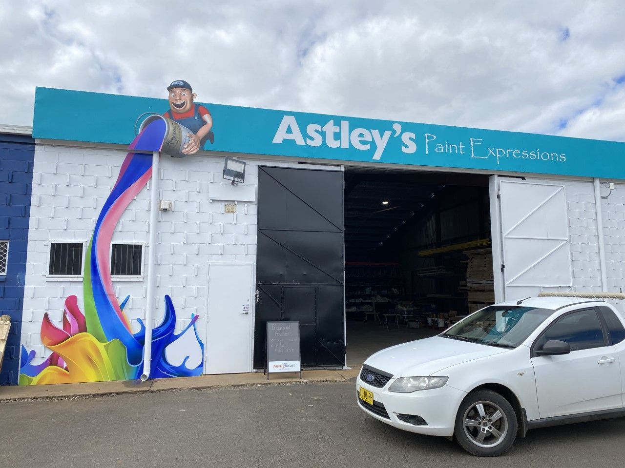 Astley's paint store with a mural of a man pouring colorful paint. White car parked outside.