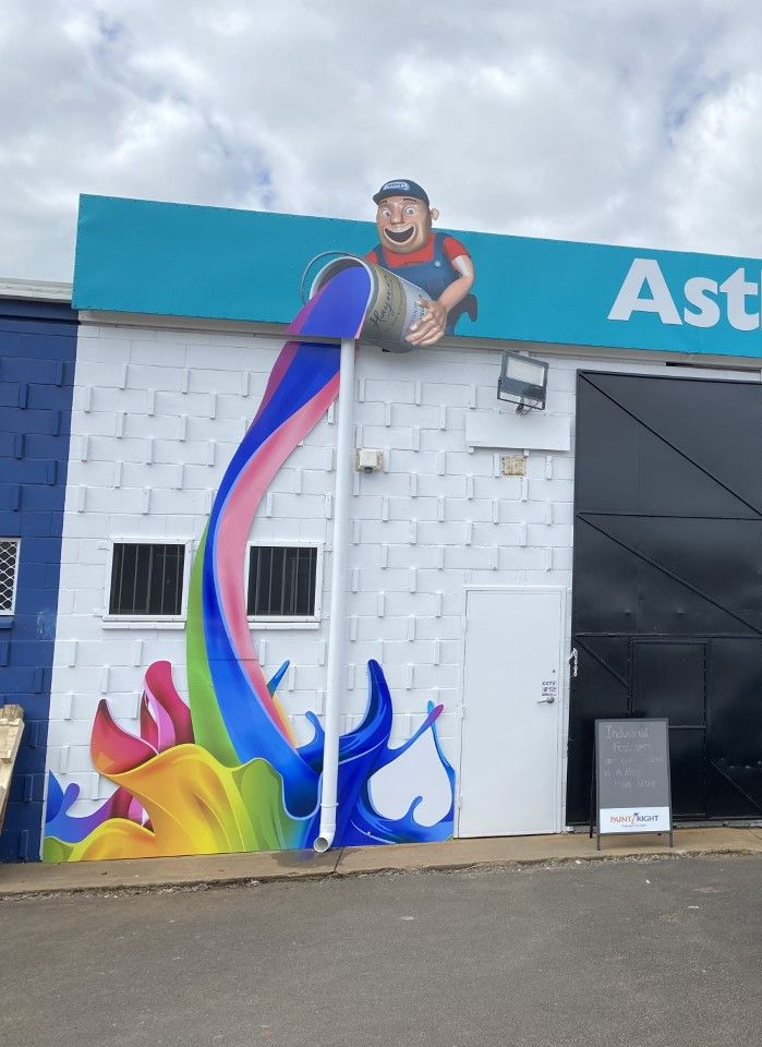 Mural of a man pouring colorful paint from a bucket down the side of a building; building is white brick with a black door.