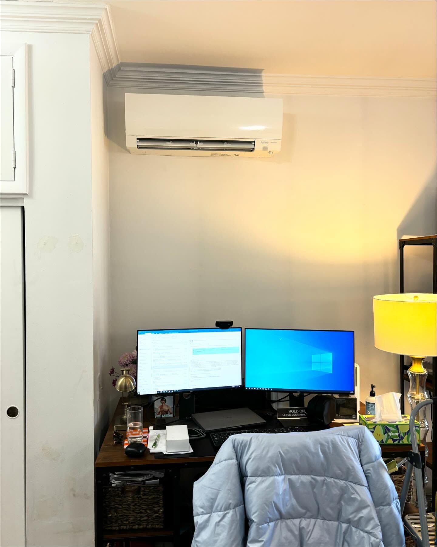 Home office desk with dual monitors, chair, lamp, and wall-mounted air conditioner. Home office desk with dual monitors, chair, lamp, and wall-mounted air conditioner.