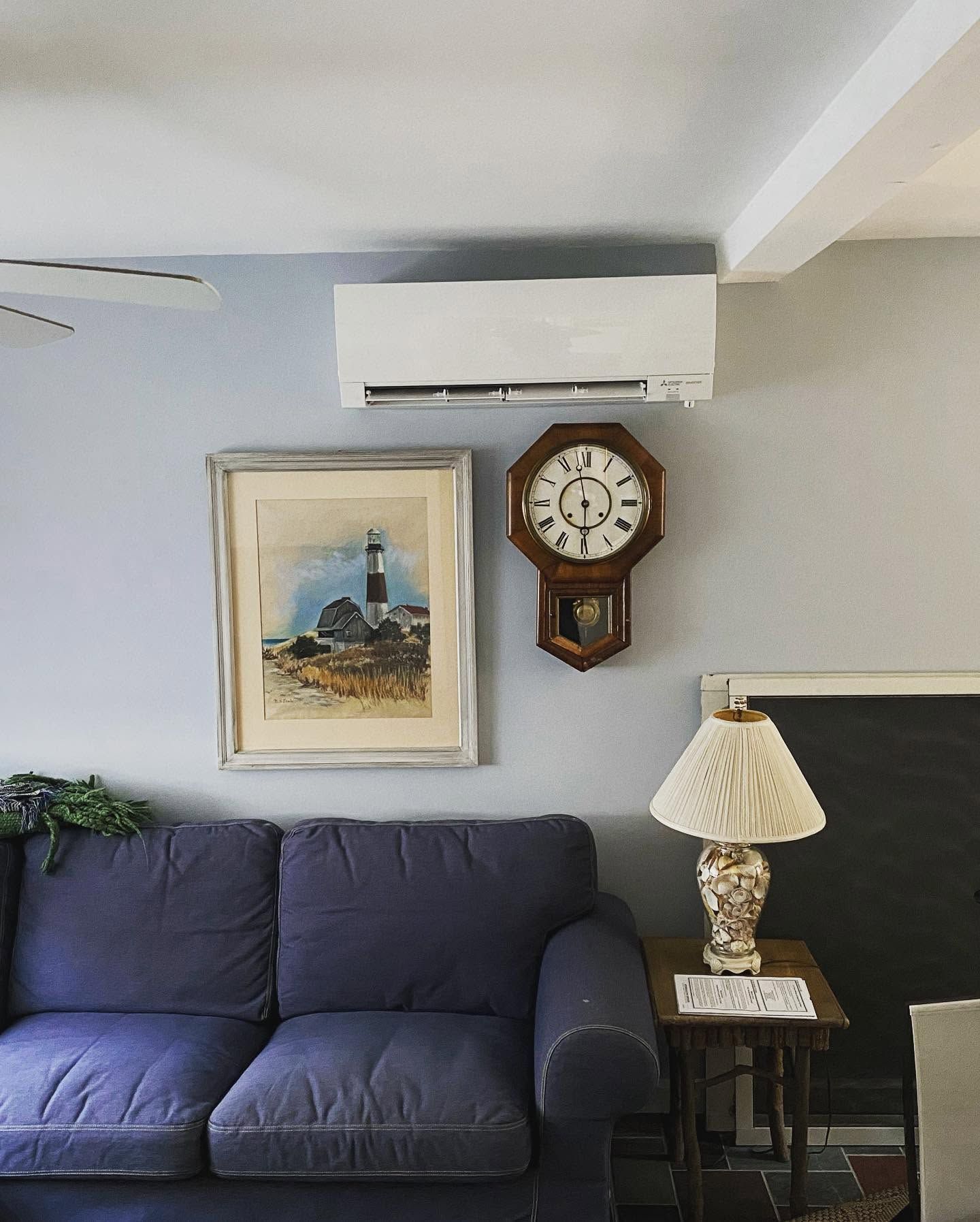 Cozy living room with blue sofa, wall clock, framed painting, table lamp, and wall-mounted air conditioner. Cozy living room with blue sofa, wall clock, framed painting, table lamp, and wall-mounted air conditioner.