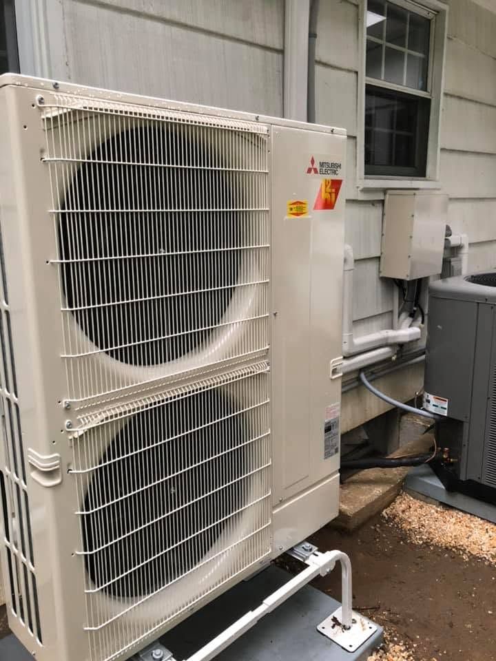 Outdoor HVAC condenser unit beside a building wall with pipes and vents, white housing and metal grille. Outdoor HVAC condenser unit beside a building wall with pipes and vents, white housing and metal grille.