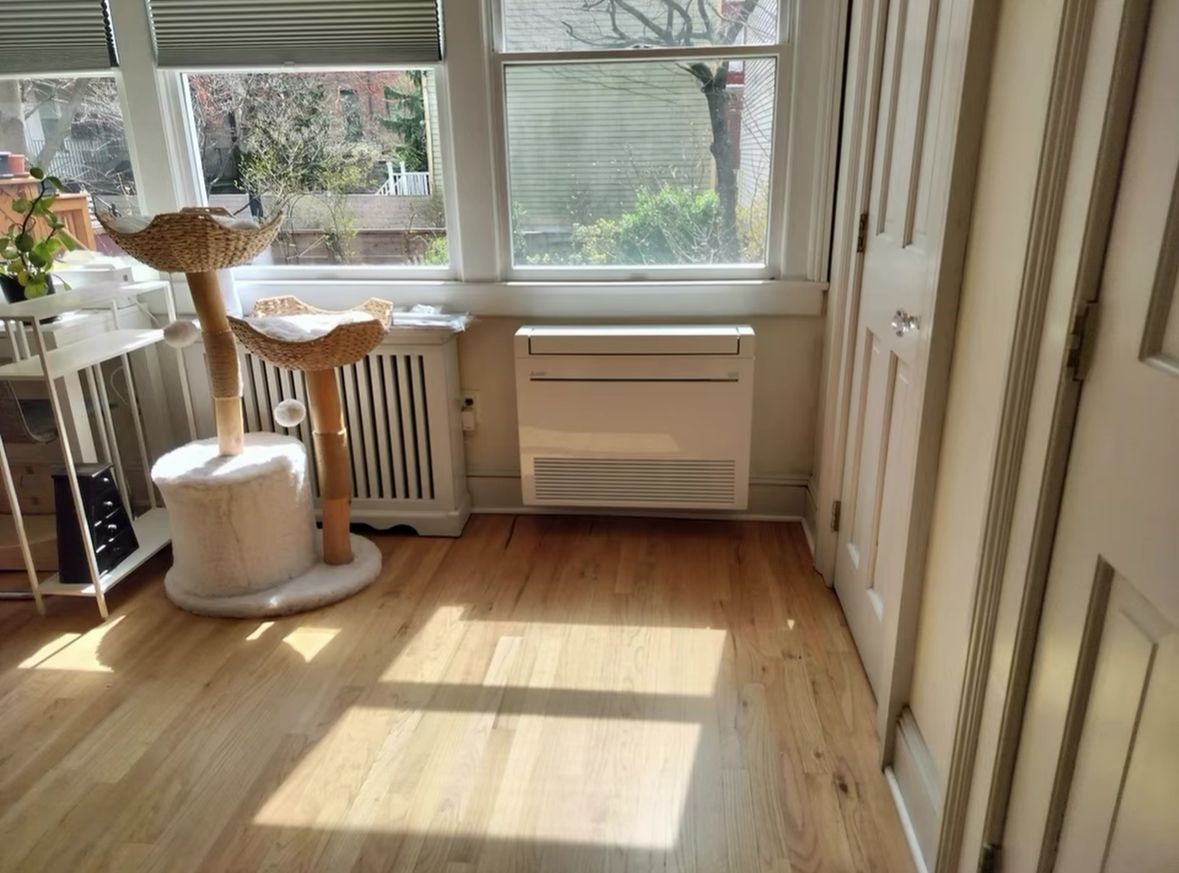 Bright sunlit room with wooden floor, window, radiator, white cat tree, and door on the right. Bright sunlit room with wooden floor, window, radiator, white cat tree, and door on the right.