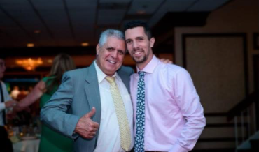 Two men smiling and posing together indoors, one in a green suit giving a thumbs-up Two men smiling and posing together indoors, one in a green suit giving a thumbs-up