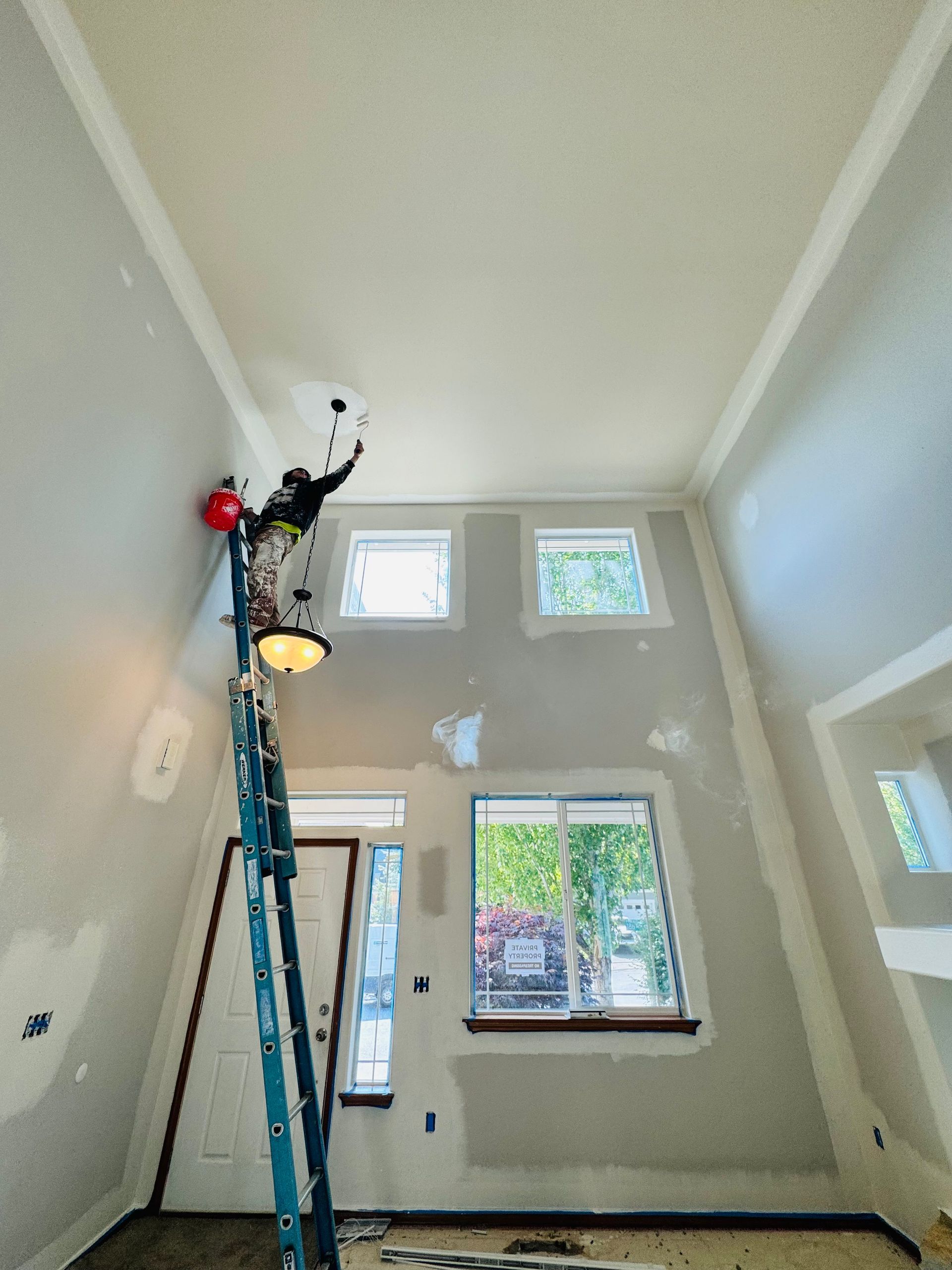 Person on ladder painting high wall in a room with windows and a doorway.