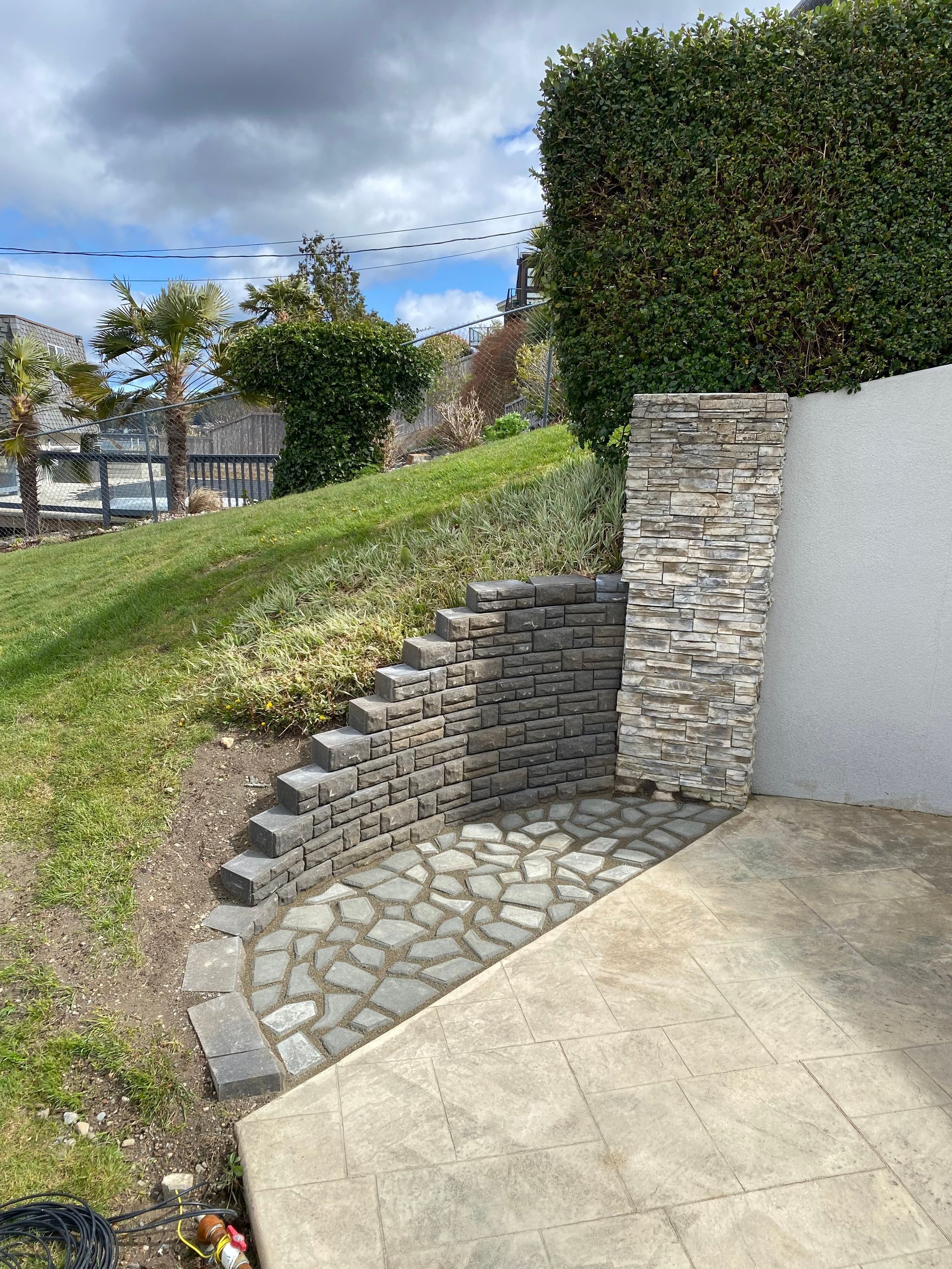 A stone retaining wall with steps and a small patio on a grassy slope next to a concrete wall.
