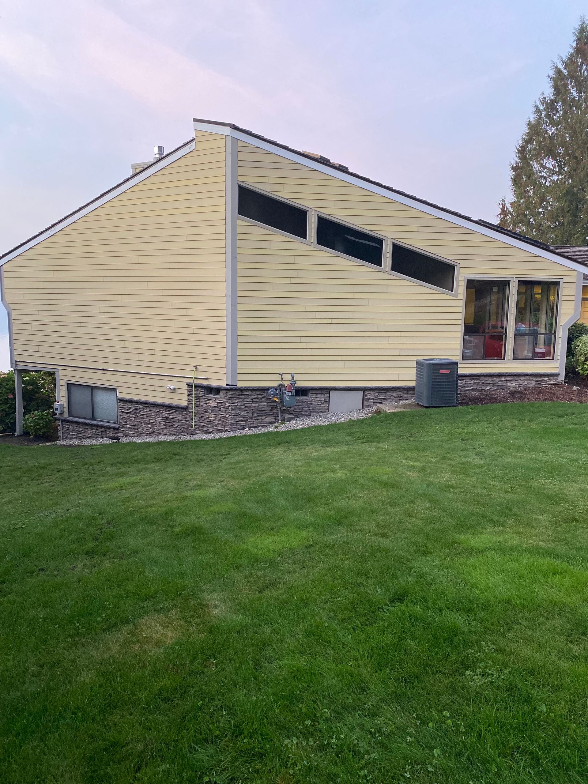 Yellow-sided house with rock foundation, set on a green lawn, with angled windows.