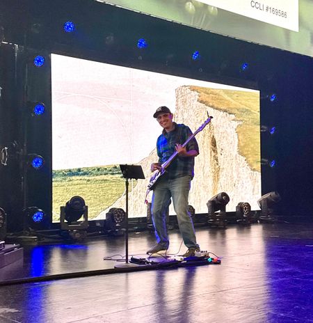 A person in a cap and plaid shirt plays a guitar on a stage in front of a large screen displaying a cliffside landscape.