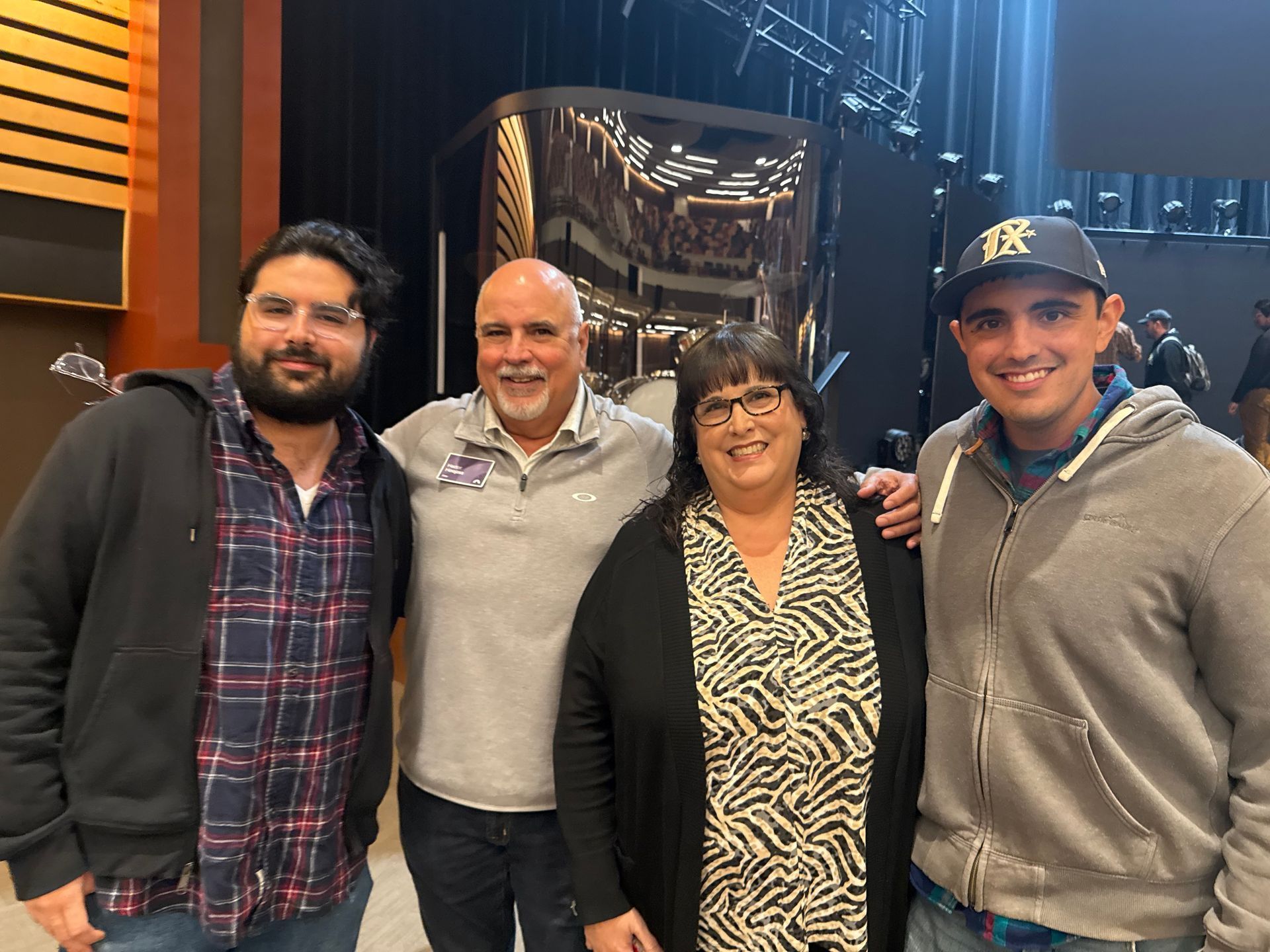 Four people posing together indoors, smiling. A metallic structure is visible in the background.