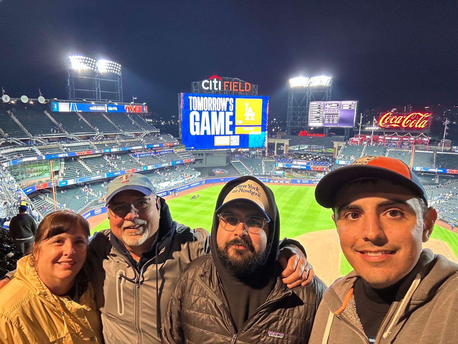 Four people smiling at a baseball game at night; Citi Field scoreboard visible.