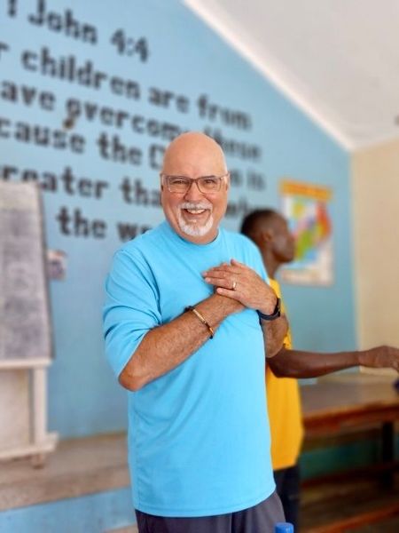 A man in a light blue long-sleeved shirt holds his hands over his heart and smiles, with text on a blue wall behind him.