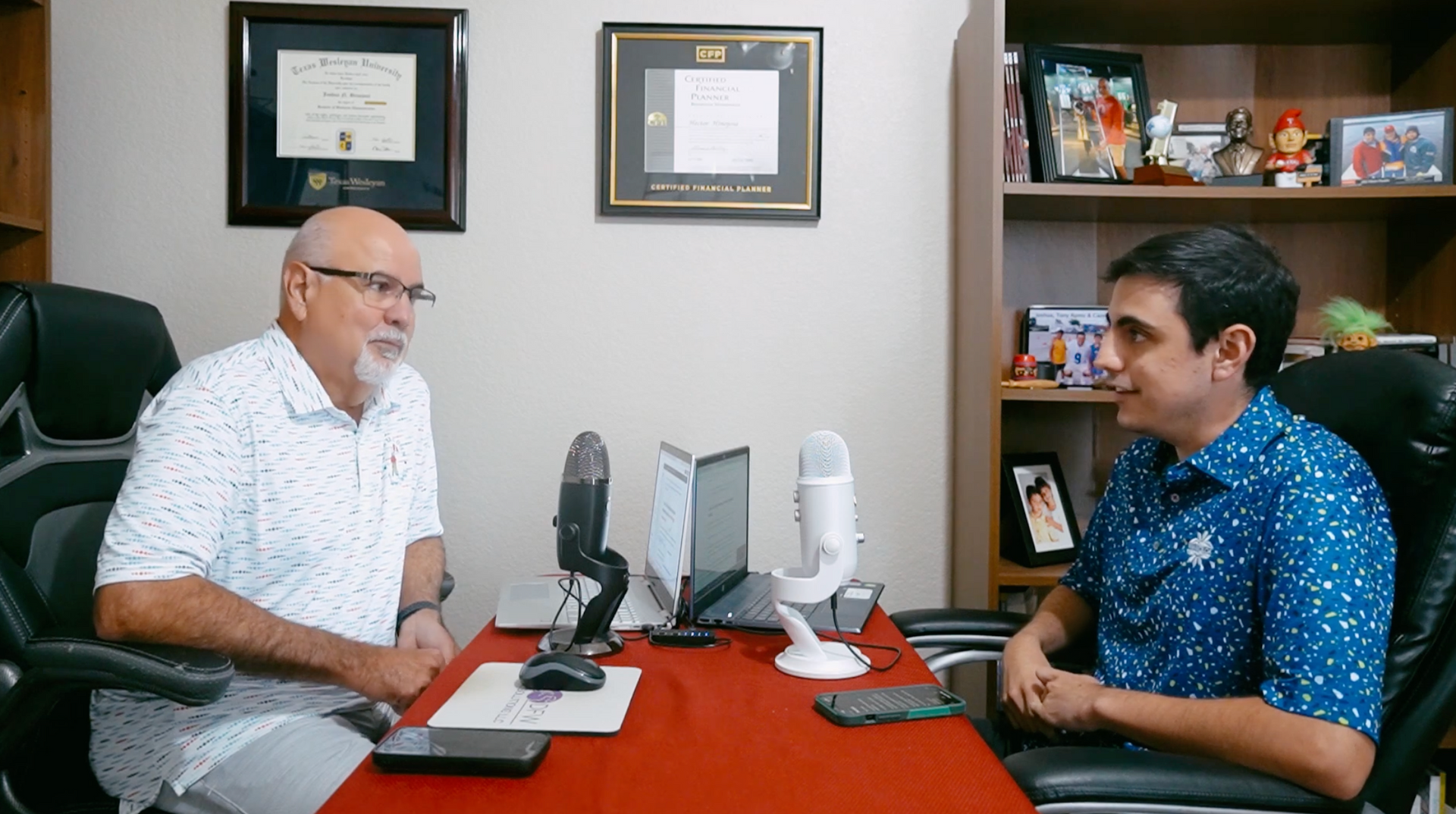 Two men at a desk with microphones, a laptop, and documents, likely an interview.