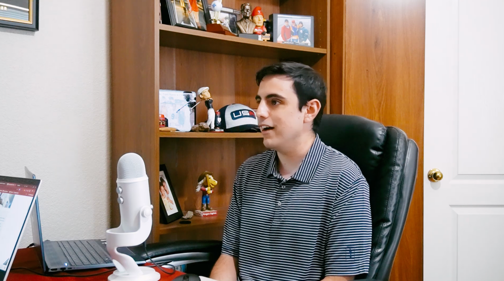 Man in a striped shirt sits at desk, speaking into microphone. Bookshelf and laptop are visible in the background.