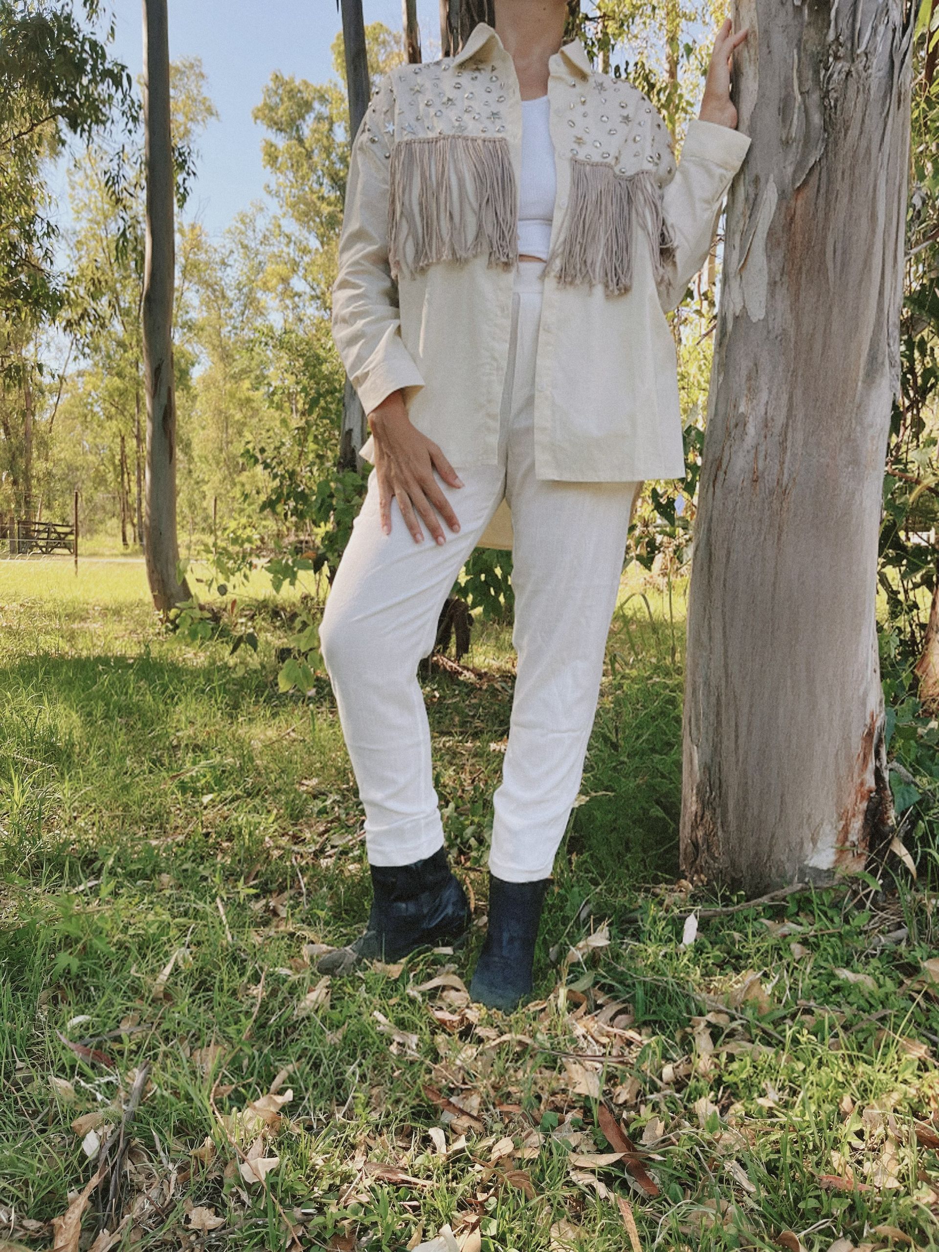 Una mujer con una chaqueta blanca y pantalones blancos está parada junto a un árbol.