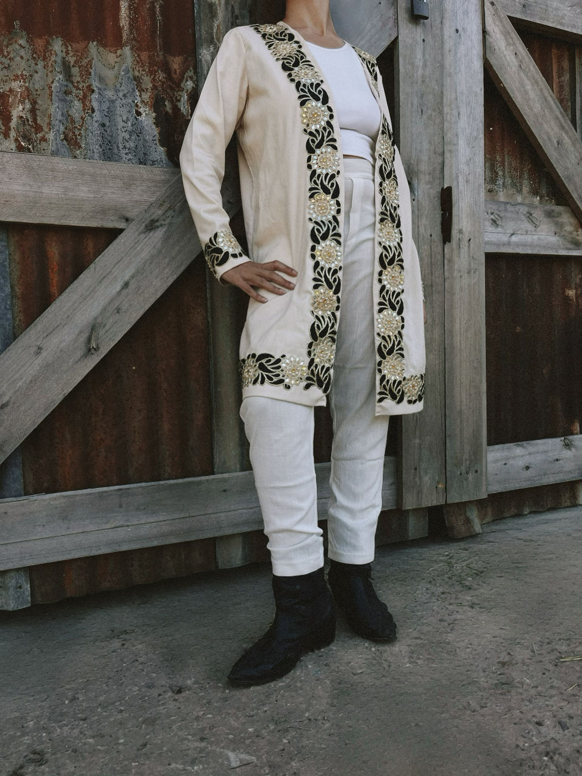 Una mujer vestida con una chaqueta blanca y pantalones blancos está parada frente a una valla de madera.