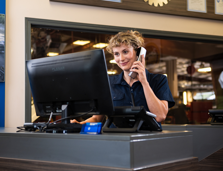 Woman in a blue shirt at a desk, talking on a phone, looking at a computer screen | AUTOPRO Oakville