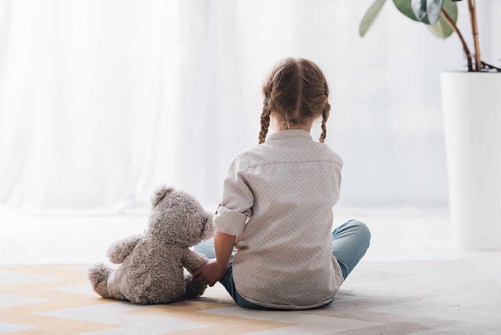 Sad little child sitting on ground with teddy bear and plant for interior design on the right hand side of photo