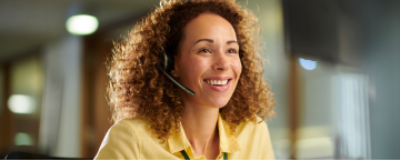 A woman is wearing a headset and smiling while sitting at a desk.