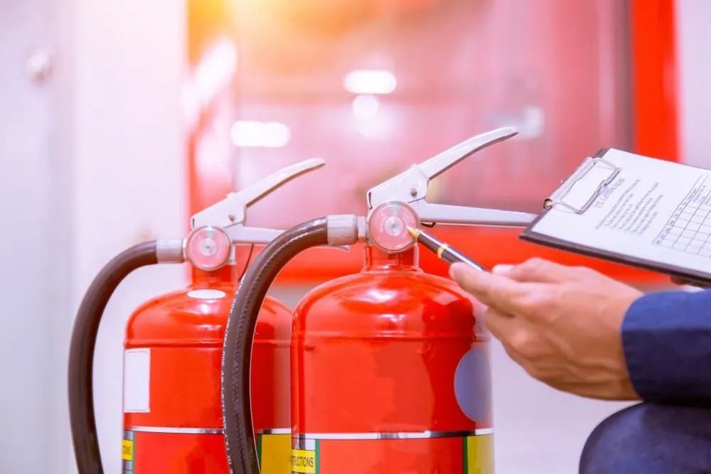 Person Inspecting Two Red Fire Extinguishers With a Clipboard — FNQ Smoke Alarms & Maintenance in Atherton, QLD