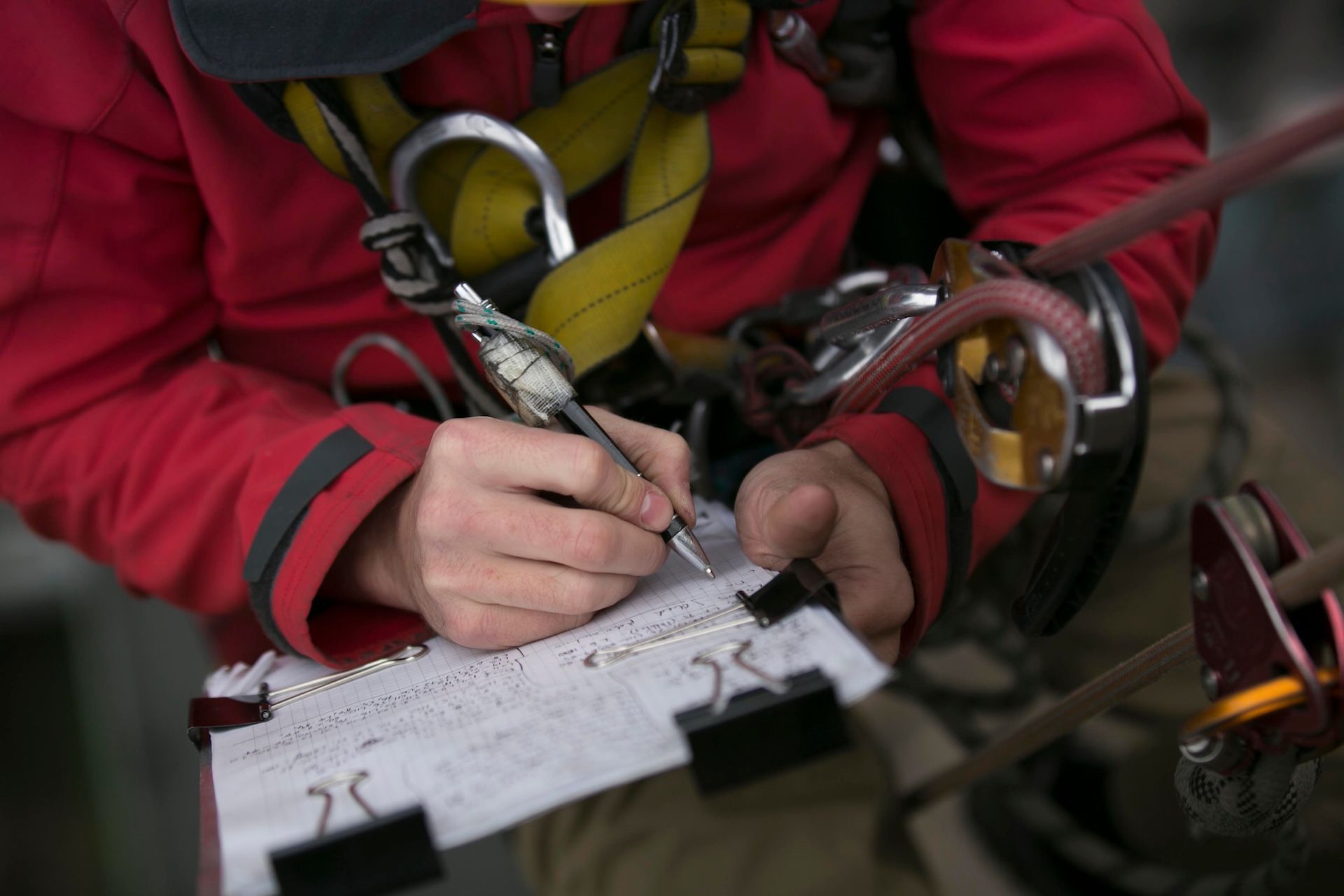 Person in Red Jumpsuit Writing on Clipboard, Wearing Safety Harness — FNQ Smoke Alarms & Maintenance in Yungaburra, QLD
