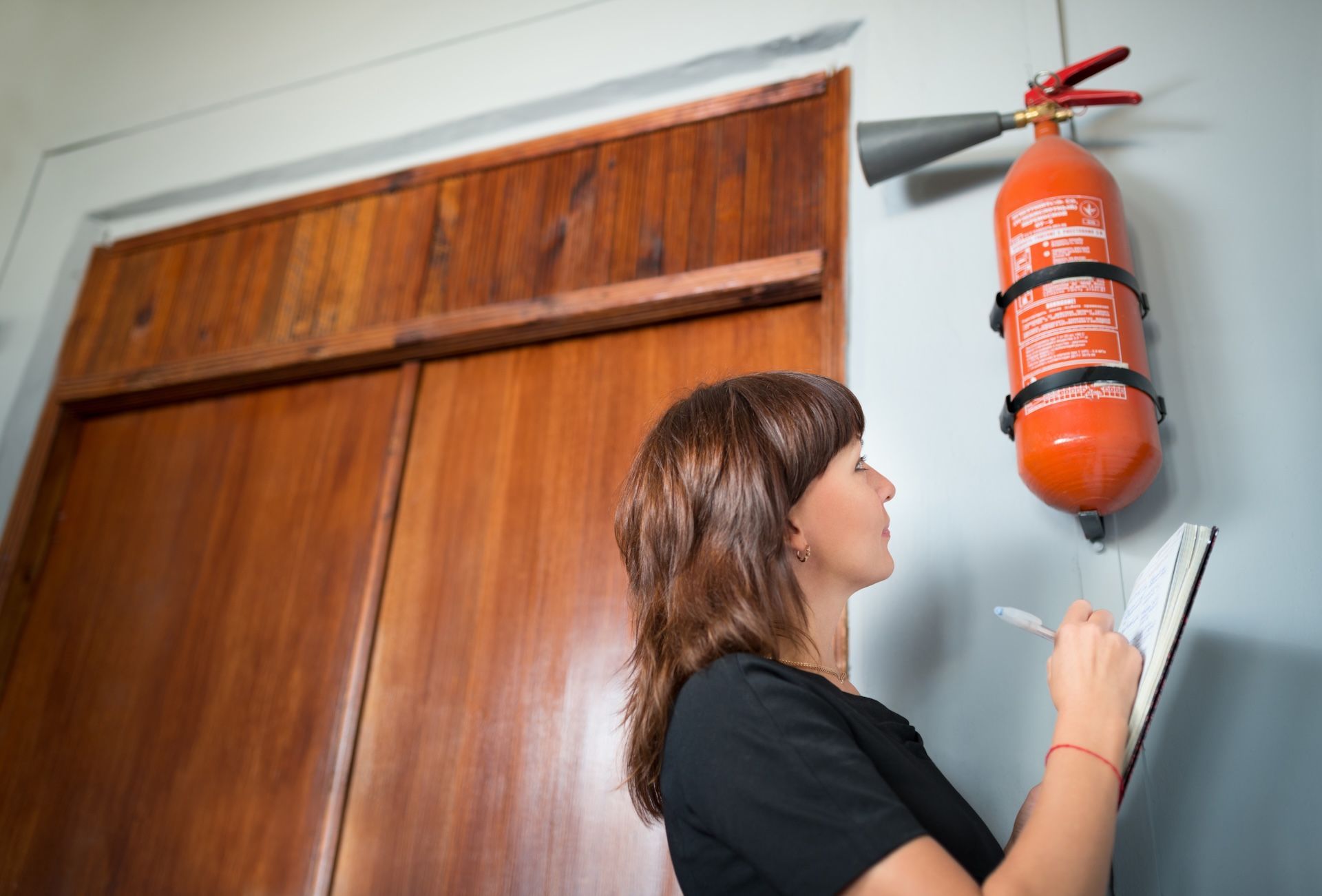 Woman Inspecting a Fire Extinguisher on a Wall — FNQ Smoke Alarms & Maintenance in Malanda, QLD