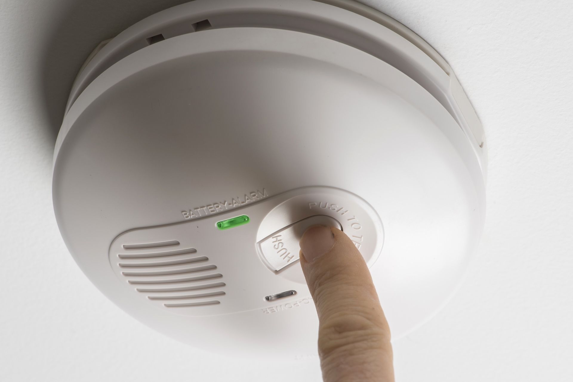 A Finger Presses a Button on a White Smoke Detector Mounted — FNQ Smoke Alarms & Maintenance in Port Douglas, QLD