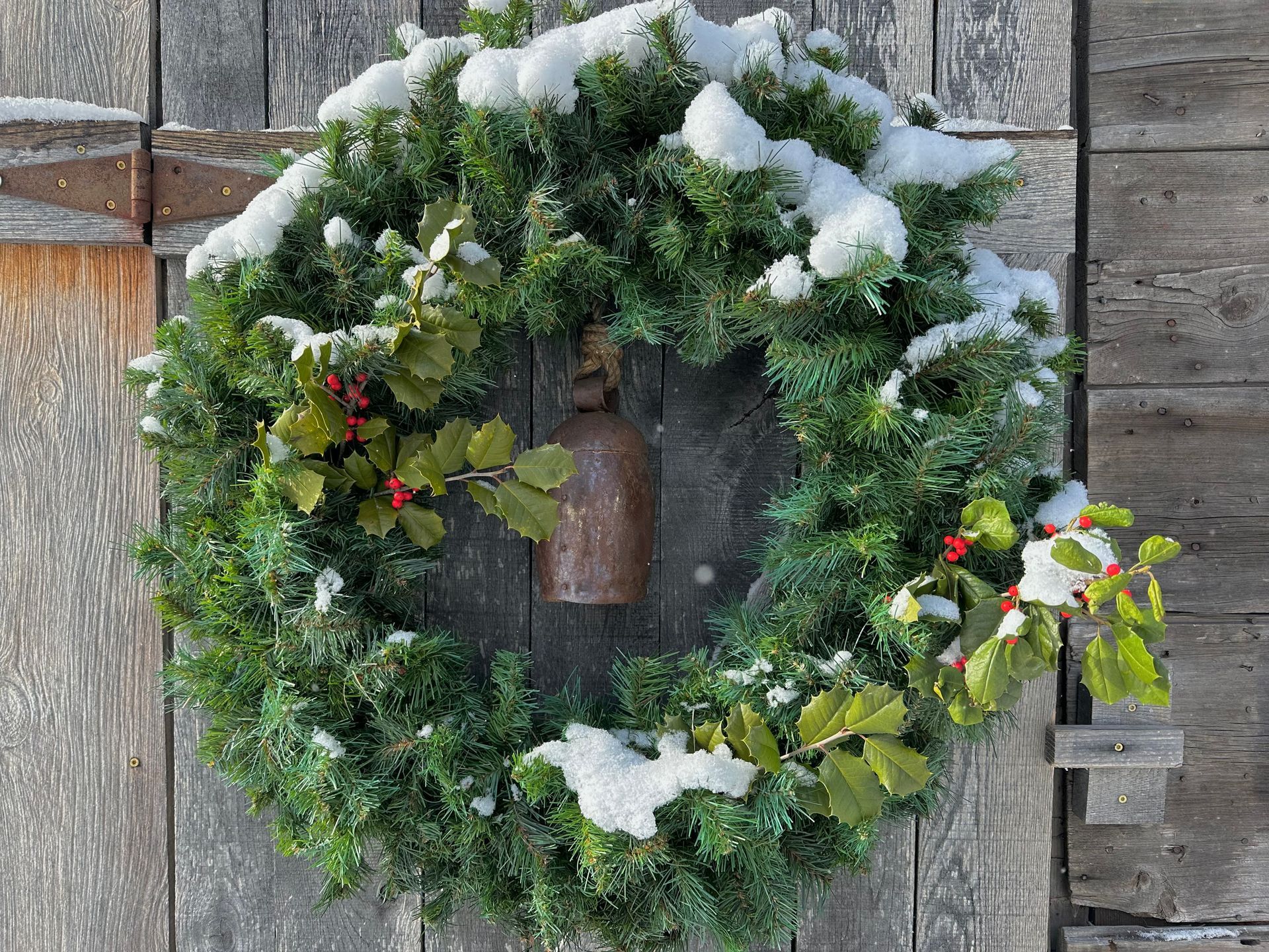 Snow-covered evergreen wreath with holly and a small bell, hanging on a weathered wooden door.