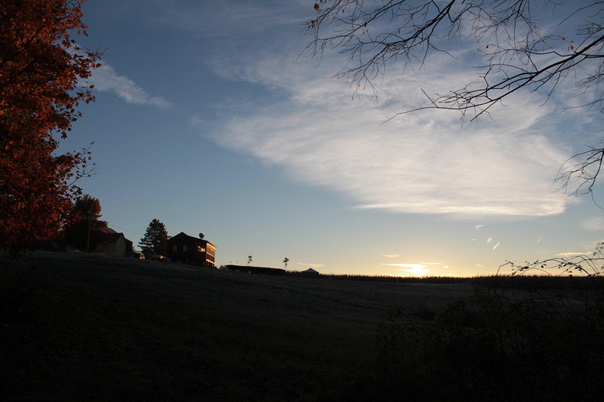 Sunset over a dark landscape with a house silhouetted on a hill. Autumn foliage and bare branches frame the sky.