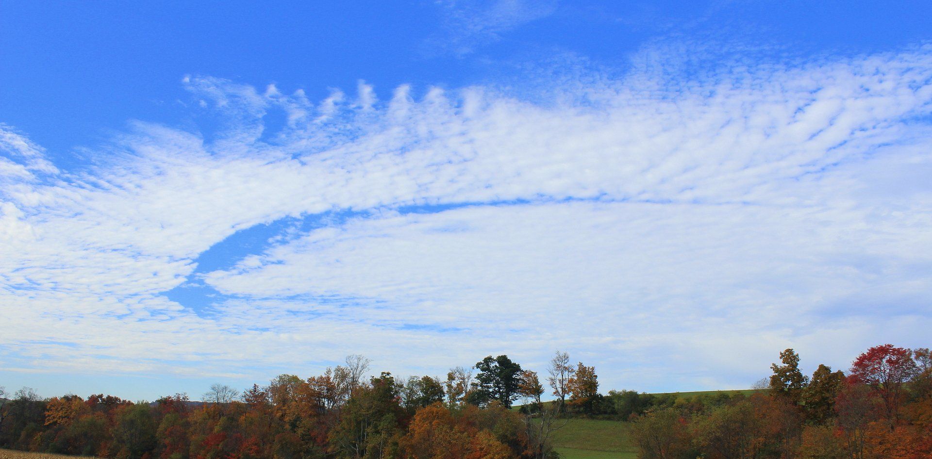 Bright blue sky with streaky white clouds forming an arc over a green hill with fall foliage.
