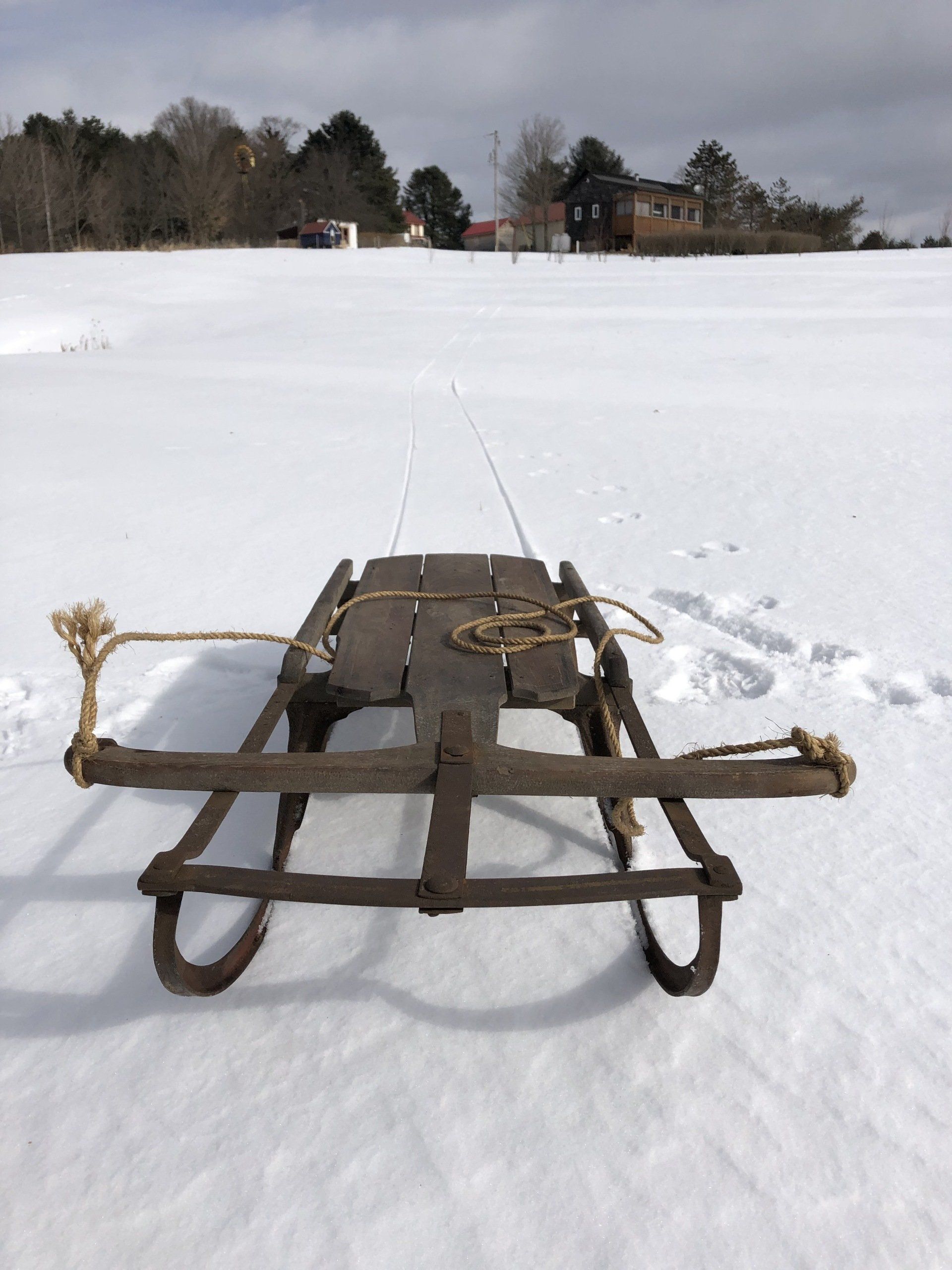 Antique wooden sled on snow-covered field, rope handles, trees and buildings in the background.