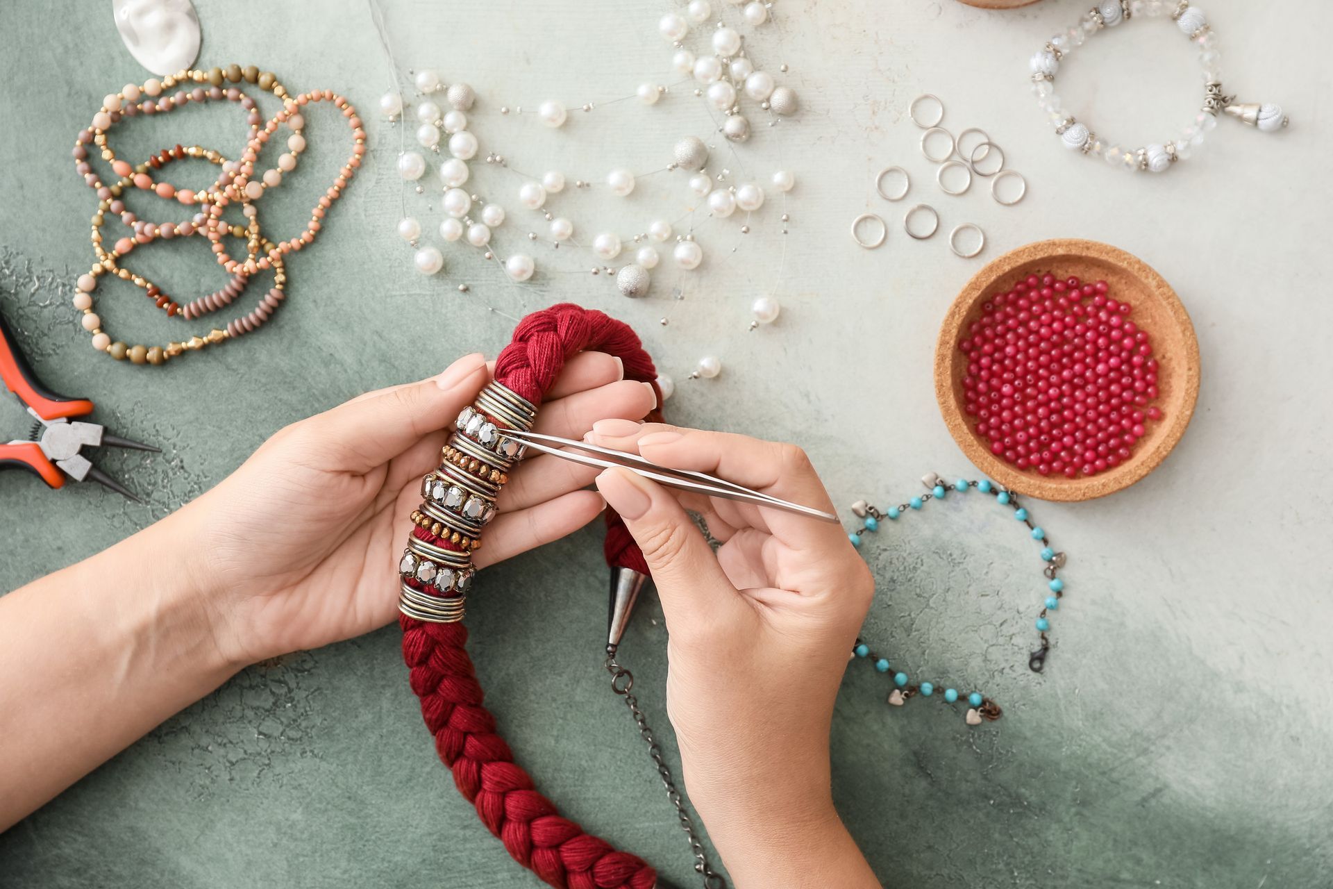 Hands beading a red necklace with jewelry-making tools and supplies on a table.