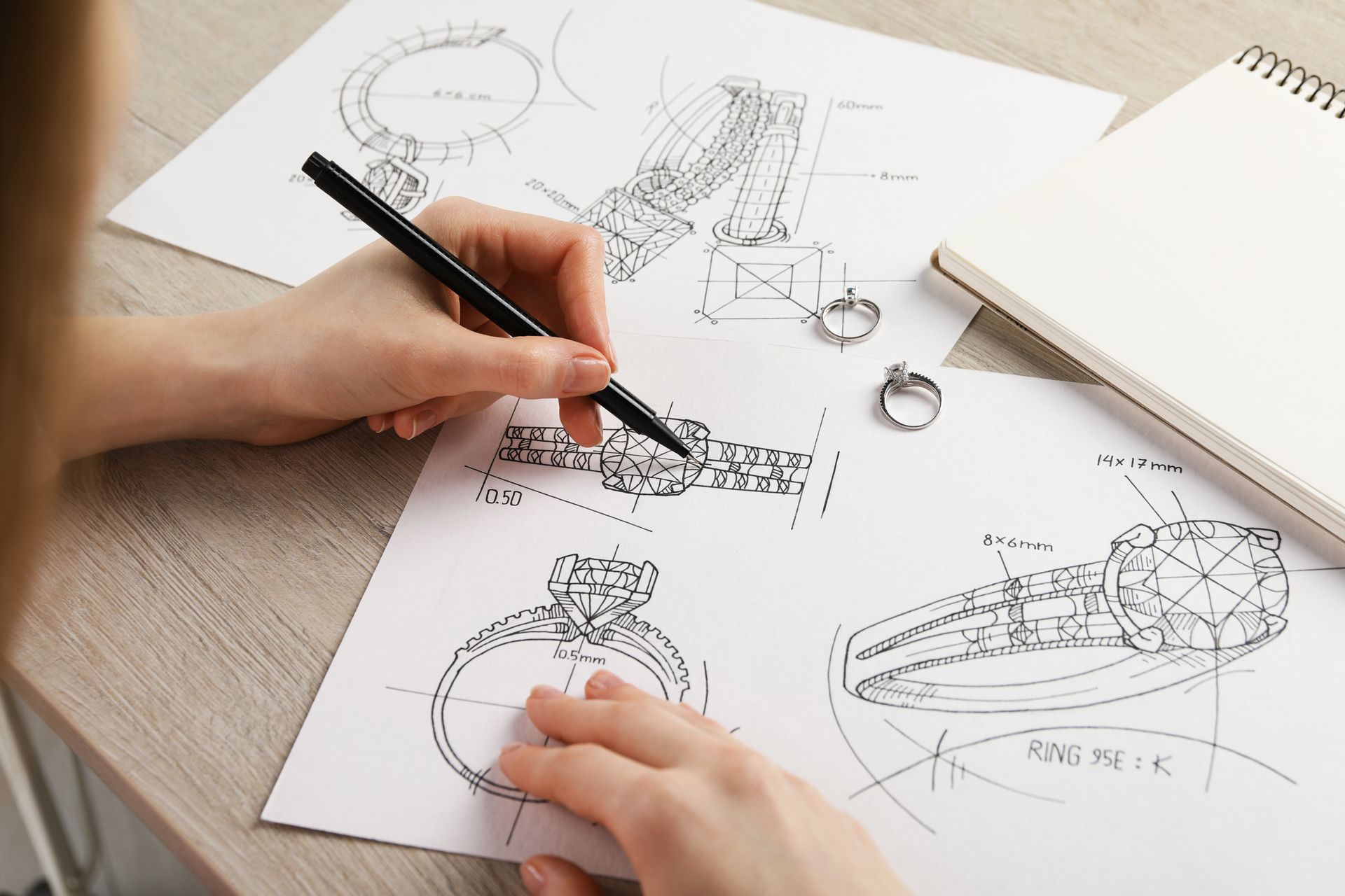 Close-up of a jeweller sketching a ring on paper at a wooden table, showcasing jewellery design.