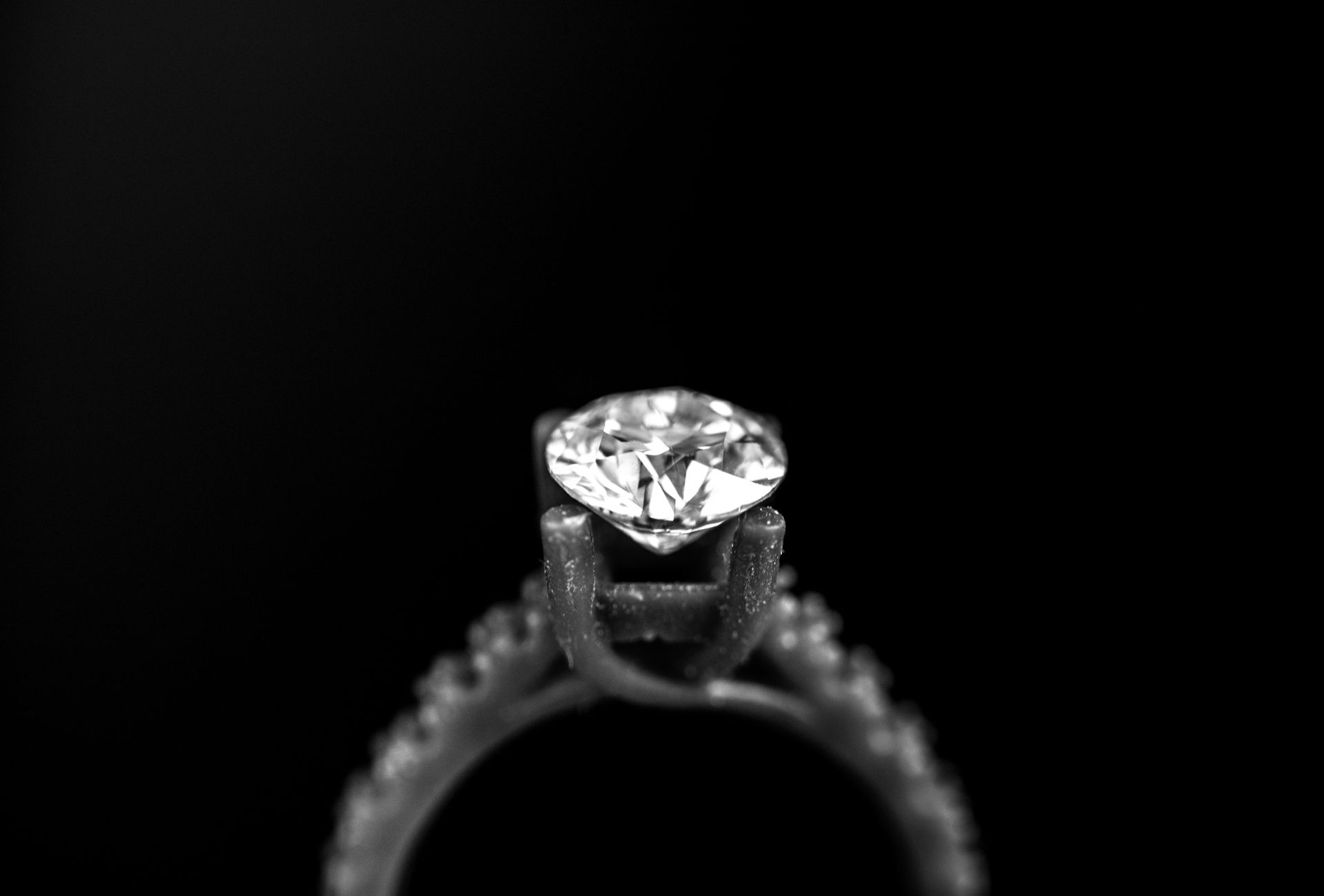 Diamond engagement ring, round-cut diamond center stone, set on a pavé band, against a black background.