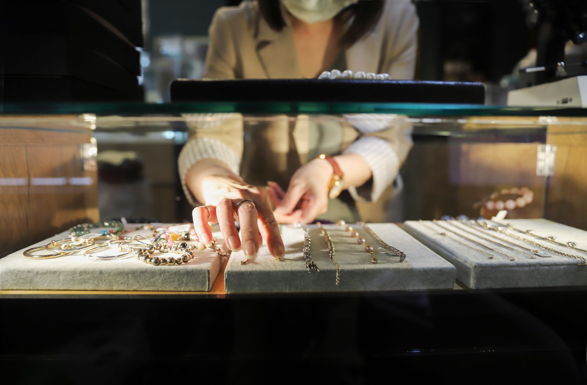 Woman wearing a face mask arranging jewelry inside a display case. Beige blazer, jewelry store setting.
