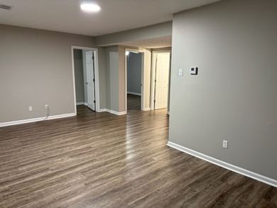 Empty, bright room with white walls, window, radiator, and light wood floors. Opening to a second room.