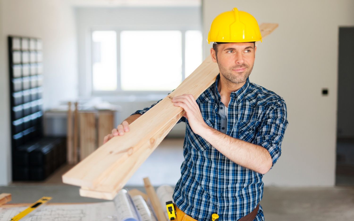 A man wearing a hard hat is carrying a piece of wood.