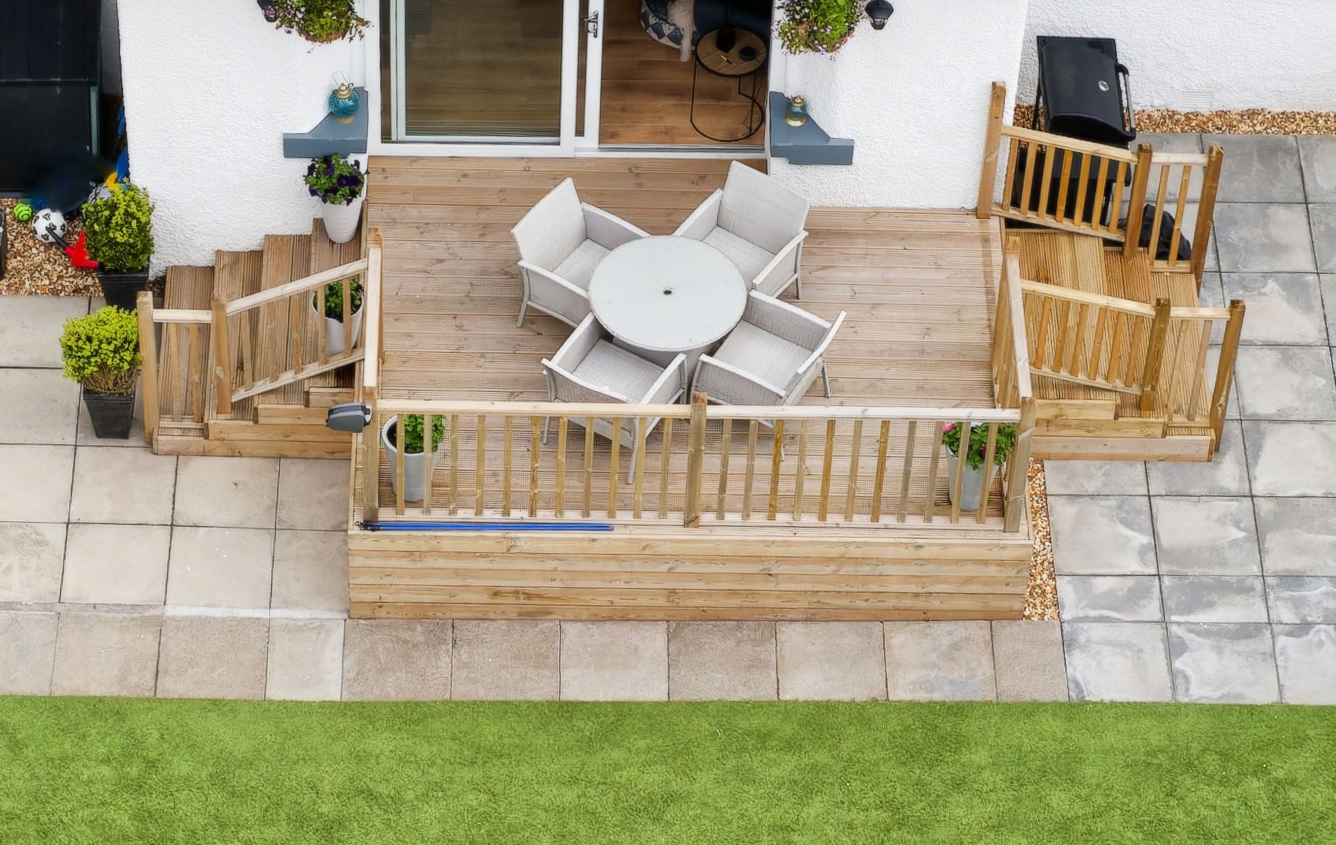 A wooden patio deck with a table and four chairs, featuring railing, side steps, and potted plants on a stone courtyard.