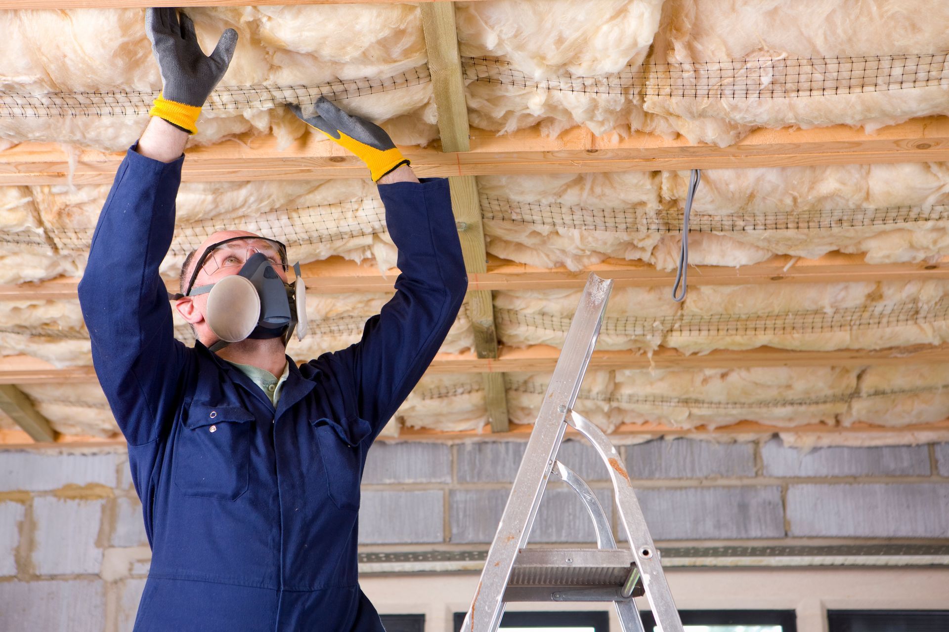Person in blue overalls and respirator installing insulation in a building's ceiling, standing on a ladder.