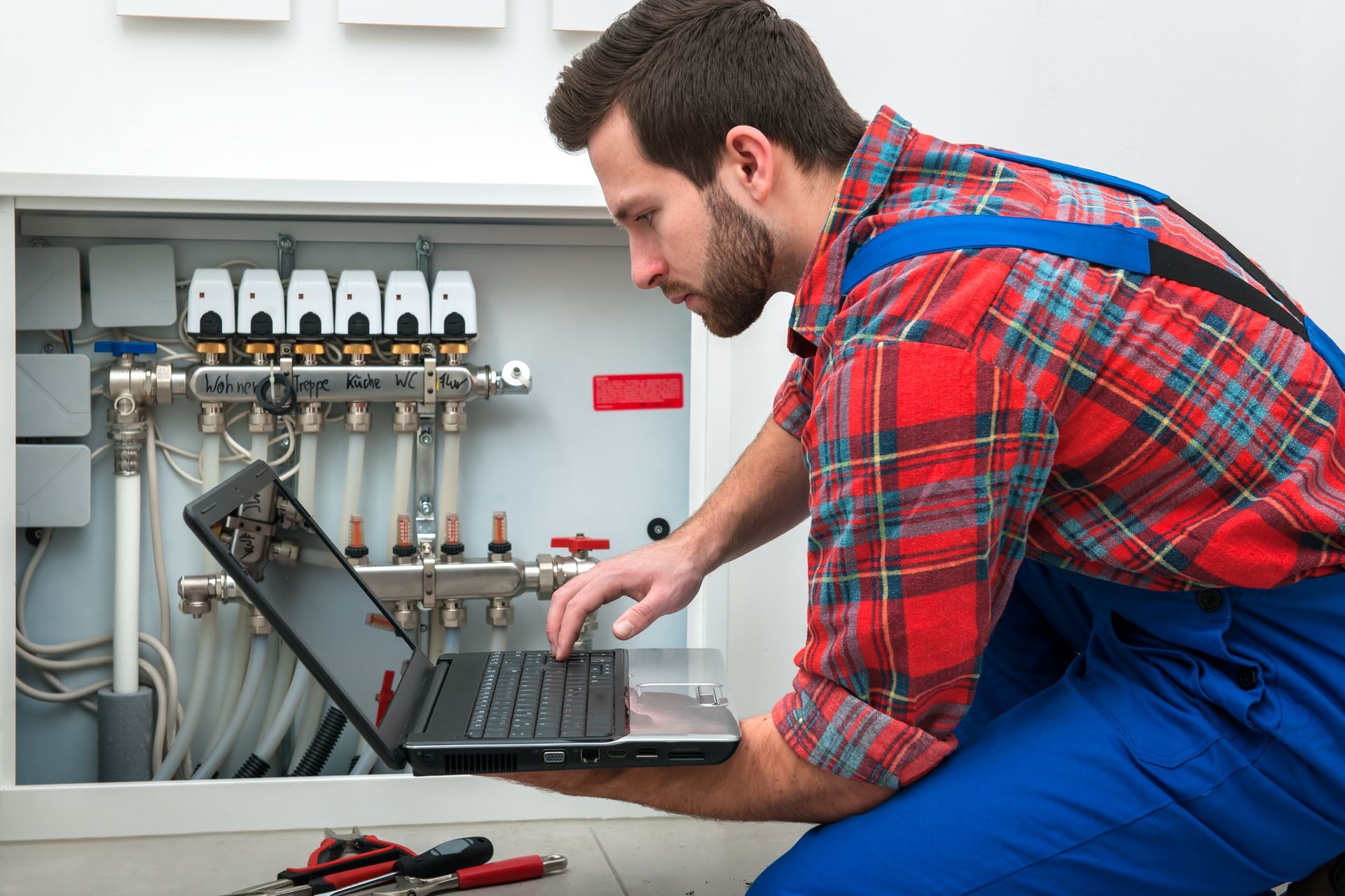 A technician in work coveralls uses a laptop to program a wall-mounted heating manifold system.