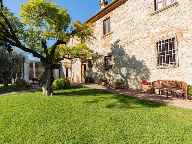 Una casa colonica soleggiata con muri in pietra, un prato verde, un grande albero frondoso e una panchina di legno su un patio piastrellato.