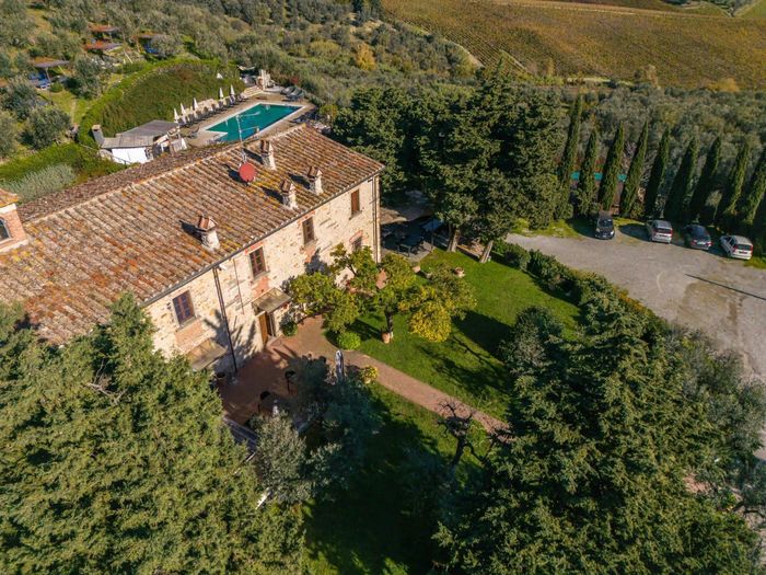 Veduta aerea di una villa rustica in pietra con tetto di tegole, dotata di piscina adiacente e area parcheggio in ghiaia.