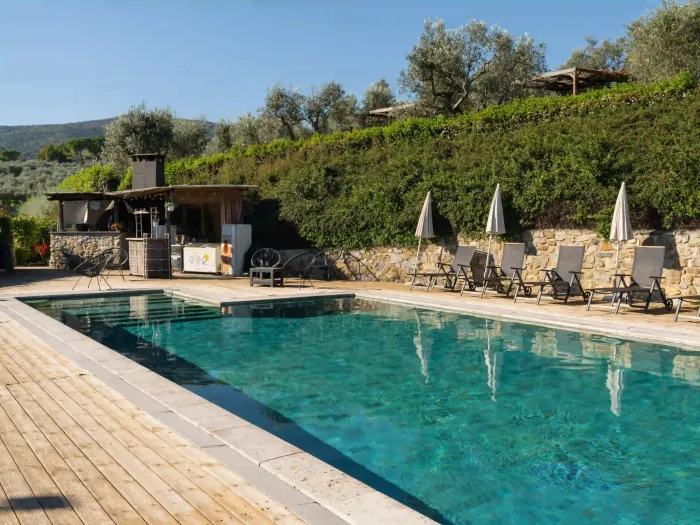 Una piscina rettangolare con acqua cristallina color turchese, circondata da una terrazza in legno, lettini prendisole e con lo sfondo di una collina.