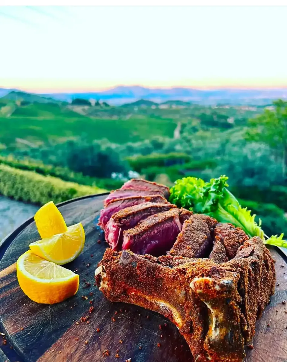 Una bistecca T-bone affettata e condita, servita su un tagliere di legno con spicchi di limone e verdure, con vista su un paesaggio suggestivo.
