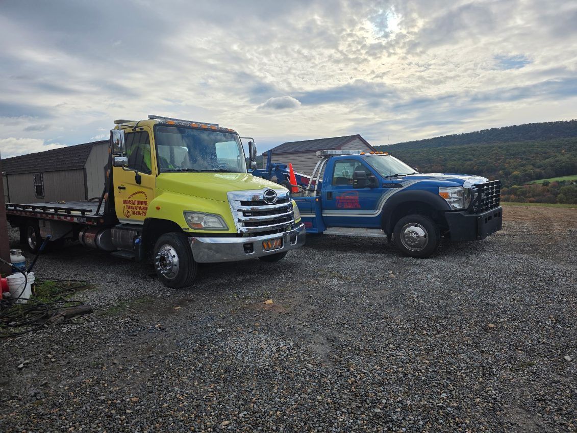 Two tow trucks, yellow and blue, parked on gravel outside a building under a cloudy sky.